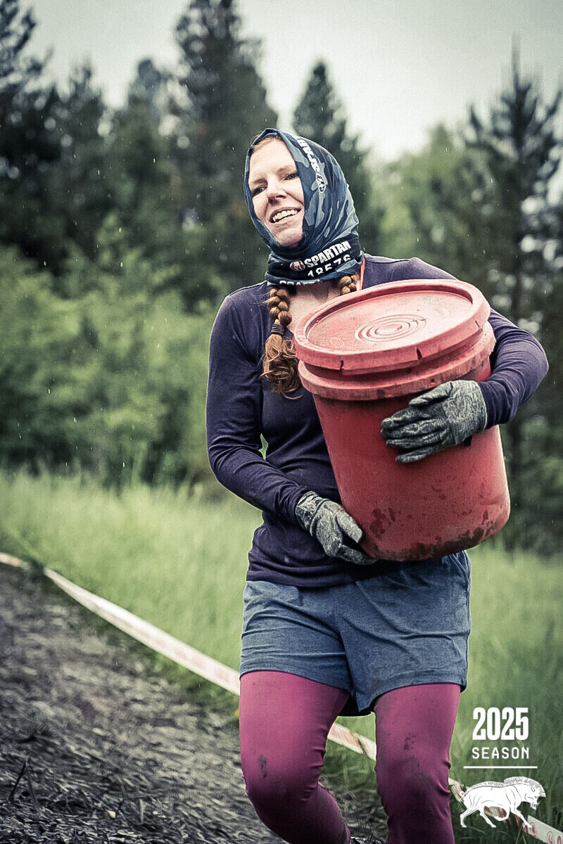 Jess smiling outdoors during an endurance race, carrying a red bucket.